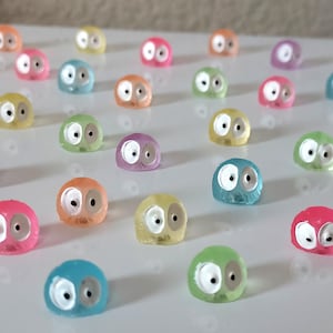 May include: A collection of colorful, translucent, small, round figurines with googly eyes. The figurines are arranged in rows on a white surface.