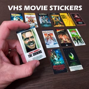 May include: A collection of VHS movie stickers featuring iconic films like "The Shining," "Back to the Future," and "Kill Bill." The stickers showcase movie posters with vibrant colors and classic film titles. The text "VHS MOVIE STICKERS" is at the top.
