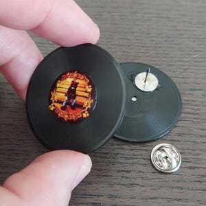 May include: Black vinyl record pin with a colorful image of a bear in the center. The pin has a silver backing and a pin closure. The image is on a wooden surface.