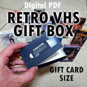 Puede incluir: Una caja de regalo de papel en miniatura diseñada como una cinta VHS negra, con una etiqueta de El silencio de los corderos. La caja se sostiene abierta para mostrar un interior blanco, rodeada de otras fundas de películas VHS en miniatura.