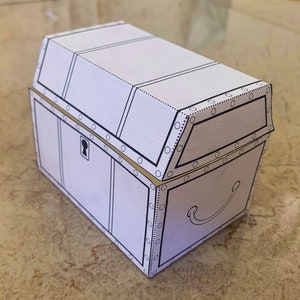 Puede incluir: Un cofre del tesoro de cart&oacute;n blanco con un borde dorado y una cerradura. El cofre est&aacute; cerrado y tiene una manija en el lateral.