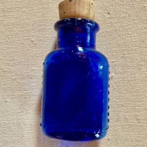 May include: A small, cobalt blue glass bottle with a cork stopper. The bottle has a rounded body and a narrow neck. The surface of the bottle has a slightly textured appearance. The cork is a light brown colour.
