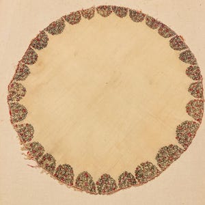 May include: A round, off-white textile with a scalloped edge. The edge is decorated with a repeating pattern of small, floral-like motifs in red, black, and green. The center is plain and unadorned.