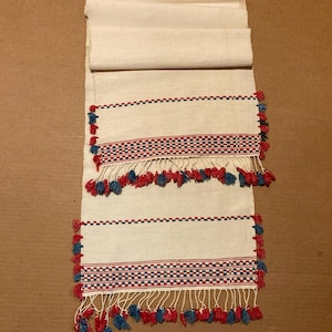 Può includere: Due runner da tavolo in lino bianco con motivi intrecciati rossi e blu e frange a nappine. Le nappine sono rosse e blu.