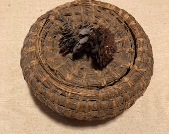 Puede incluir: Una cesta redonda tejida con tapa, hecha de materiales naturales marrones. La tapa está decorada con un pequeño grupo de piñas secas.