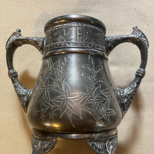 May include: A silver sugar bowl with a floral design and three feet. The bowl has two handles and a decorative band around the top.