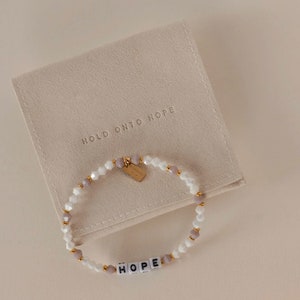 Könnte beinhalten: Ein weißes und graues Perlenarmband mit einem goldenen Anhänger, auf dem "HOPE" steht. Das Armband befindet sich in einem kleinen, beigen Beutel mit dem Aufdruck "HOLD ONTO HOPE".