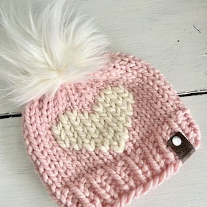 May include: Pink knitted hat with a white heart design and a white faux fur pom pom. The hat has a brown leather tag with the text "Handmade with Love" on it.