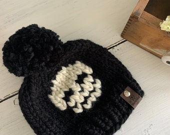 May include: Black knit beanie with a white skull design and a brown leather tag that says "Little Fox Knit". The beanie has a fluffy pom pom on top.
