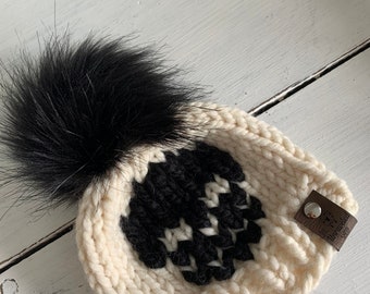 May include: White knitted beanie with a black skull design and a black faux fur pom pom. The beanie has a brown leather tag with the text "Alora Made" on it.