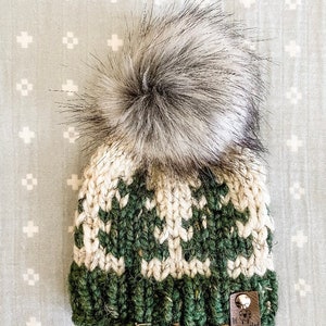 May include: A green and white knitted hat with a faux fur pom pom on top. The hat has a brown leather tag with the text "The Unraveled" on it.