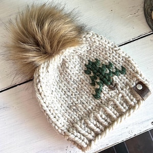 May include: A cream-colored knitted beanie with a green tree design and a brown faux fur pom pom. The beanie has a brown leather tag with the text "Handmade with Love" on it.