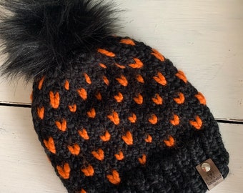May include: Black knit beanie with a black faux fur pom pom and orange heart pattern. The beanie has a brown leather tag with the text "Unraveled Sheep".
