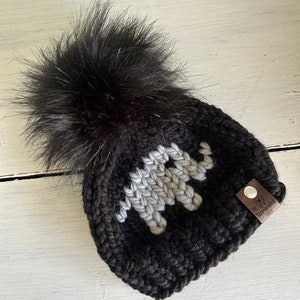 May include: Black knit beanie with a white knit elephant design and a black faux fur pom pom. The beanie has a brown leather tag with the text "The Warveled Shop".