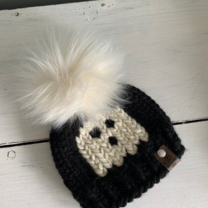 May include: Black and white knit beanie with a white pom pom and a brown leather tag that says "The Cozy Knitter".