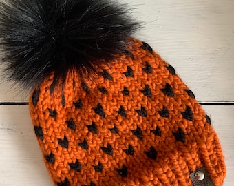 May include: Orange knit beanie with a black pom pom and black heart pattern. The beanie has a small brown leather tag with the text "Unraveled" on it.
