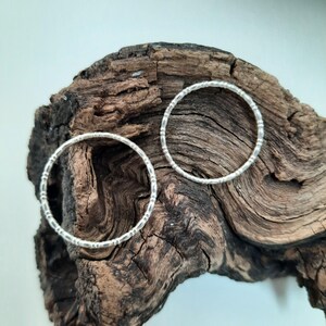 May include: Two silver hoop earrings with a textured design. The earrings are circular and feature a series of small, evenly spaced indentations around the circumference. They are displayed on a piece of dark, textured wood.