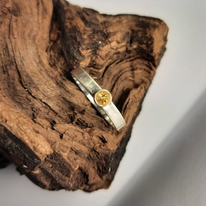 Op de afbeelding: Een zilveren ring met een kleine, ronde, gele edelsteen in het midden. De ring ligt op een stuk bruin hout.