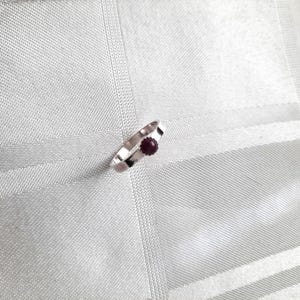 Op de afbeelding: Een zilveren ring met een kleine, ronde, donkerrode edelsteen. De ring heeft een eenvoudig, modern ontwerp met een licht getextureerde band. De edelsteen is gezet in een bezel setting.