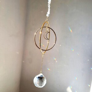 Op de afbeelding: Een hemels thema zonnevanger met een goudkleurig metalen ontwerp. Het heeft twee concentrische ringen, een maan en een heldere kristallen bol, hangend aan een ketting van heldere kralen.