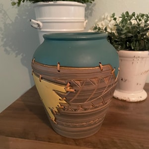 May include: A ceramic vase with a teal upper section and a brown lower body featuring carved geometric patterns. A bright yellow triangular accent piece is attached to the side with tan leather lacing that wraps around the upper rim.