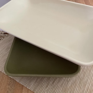 May include: Two rectangular ceramic serving platters, one cream-colored and the other olive green, are stacked on top of each other. The platters have rounded corners and are placed on a beige woven placemat.