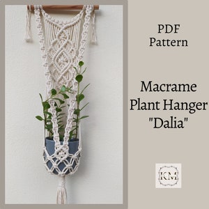 Könnte beinhalten: Ein cremefarbener Makramee-Pflanzenaufhänger, der einen grauen Topf mit grünen Pflanzen hält. Das Design umfasst eine Holzstange, komplizierte Knoten und eine Quaste. Der Text lautet "PDF Pattern" und "Macrame Plant Hanger 'Dalia'".