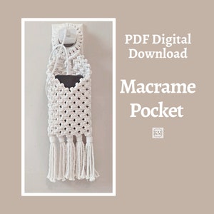 以下が含まれることがあります： 壁に掛けるようにデザインされた白いマクラメポケットで、中に電話が入っています。 ポケットは織り込まれた綿コードでできており、底にフリンジが付いています。 テキスト「PDF Digital Download Macrame Pocket」が表示されています。