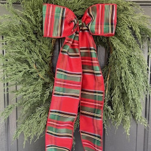 May include: A circular wreath made of dense green evergreen branches featuring a large, prominent bow. The bow is crafted from fabric with a red, green, and white plaid pattern, featuring long, flowing tails that hang down the center.