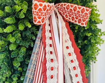 May include: A circular wreath made of dense green boxwood foliage, adorned with a large bow featuring a red and white geometric pattern. Cascading below the bow are multiple ribbons in red, white, and blue, including lace and striped textures.