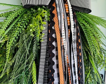 May include: A decorative door swag featuring a large black and white striped bow at the top, cascading down with multiple ribbons in orange, black, white, and gray patterns, accented by lush green fern-like foliage and small leafy sprigs.