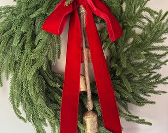 May include: A green evergreen wreath with a red velvet ribbon and hanging gold bells. The wreath is a festive decoration, perfect for the holiday season. The bells are attached with a natural rope.