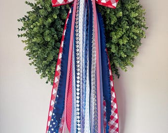 May include: A round green wreath with a red and white checkered bow. The wreath is decorated with red, white, and blue ribbons with star and stripe patterns, perfect for patriotic holidays.