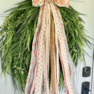 Op de afbeelding: Een groene kunstkrans met een decoratieve strik met pompoenen en linten in verschillende patronen en texturen. De linten bevatten stippen, strepen en kant, in tinten beige, oranje en grijs. Een seizoensgebonden woondecoratie-artikel.