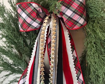 May include: A festive Christmas wreath featuring a large plaid bow with red trim. The bow is accented with gold star ribbon and cascading ribbons in red, white, black, and gold. The wreath is made of green evergreen branches.