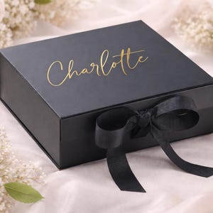 Può includere: Una scatola regalo nera con il nome "Charlotte" in oro sul coperchio. La scatola è legata con un nastro e un fiocco di raso nero. Lo sfondo presenta fiori bianchi e un tessuto bianco morbido.