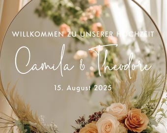 Pode incluir: Um espelho redondo com moldura dourada, decorado com flores e vegetação. O espelho mostra os nomes "Camila & Theodore" e a data "15. August 2025". O texto "WILLKOMMEN ZU UNSERER HOCHZEIT" está no topo.