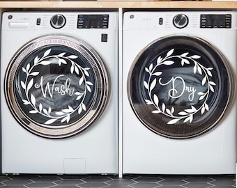 May include: White washing machine and tumble dryer with black wreath designs on the front. The washing machine says "Wash" and the tumble dryer says "Dry" in a white script font.