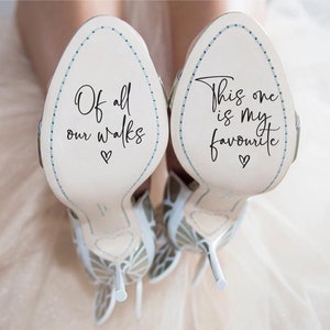 Puede incluir: Un par de zapatos de boda blancos con un diseño de mariposa azul. Las suelas de los zapatos tienen mensajes escritos a mano: "Of all our walks" y "This one is my favourite".
