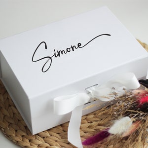 May include: A white gift box with a white ribbon and the name "Simone" written in black cursive on the top.