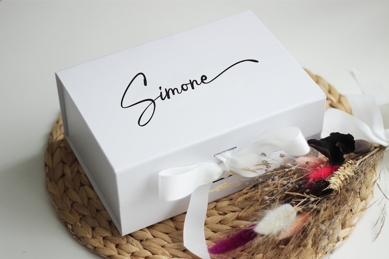 May include: A white gift box with a white ribbon and the name "Simone" written in black cursive on the top.