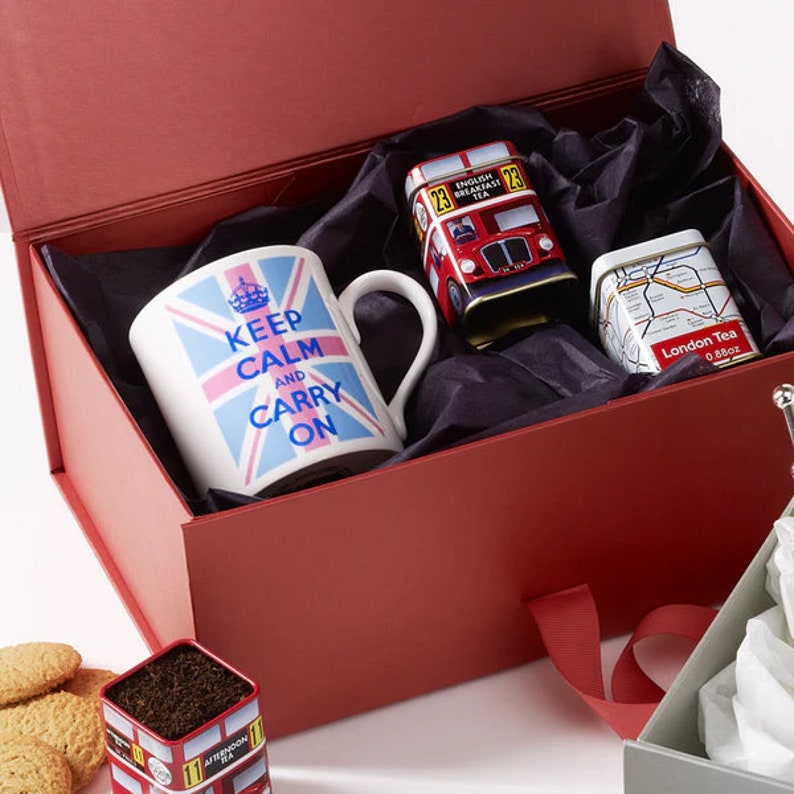 May include: A red gift box with a black and white patterned lining. The box contains a white mug with a blue and pink Union Jack flag and the text "Keep Calm and Carry On". There are also two small tin boxes, one with a red double-decker bus and the text "English Breakfast Tea" and the other with a map of London and the text "London Tea".