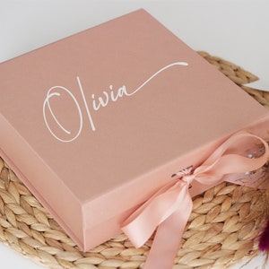 May include: A blush pink gift box with a satin ribbon tied around it. The box has the name "Olivia" written in white cursive on the top.
