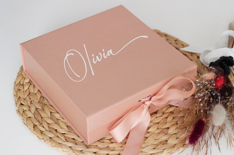 May include: A blush pink gift box with a satin ribbon tied around it. The box has the name "Olivia" written in white cursive on the top.