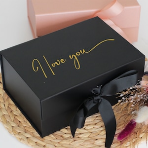 May include: A black gift box with a gold inscription that reads "I love you" and a black satin ribbon. The box is sitting on a woven straw mat.