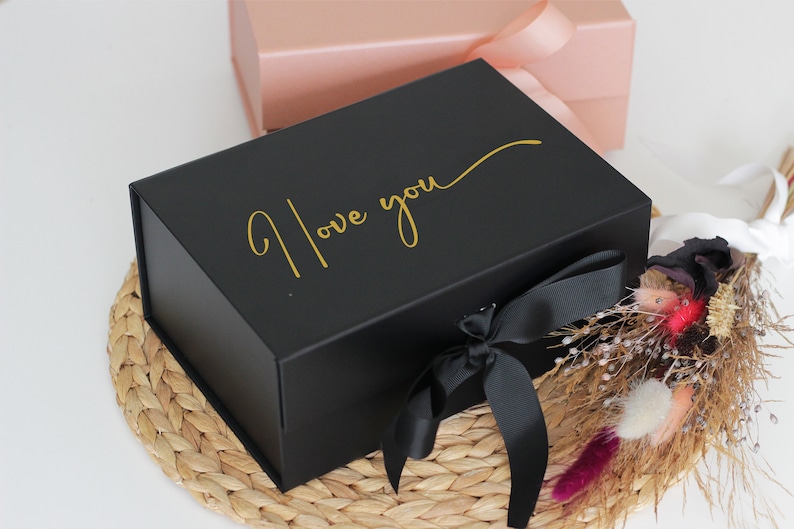 May include: A black gift box with a gold inscription that reads "I love you" and a black satin ribbon. The box is sitting on a woven straw mat.