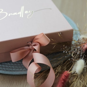 May include: A pink gift box with a gold inscription that reads "Bradley" and a blush pink satin ribbon tied in a bow.