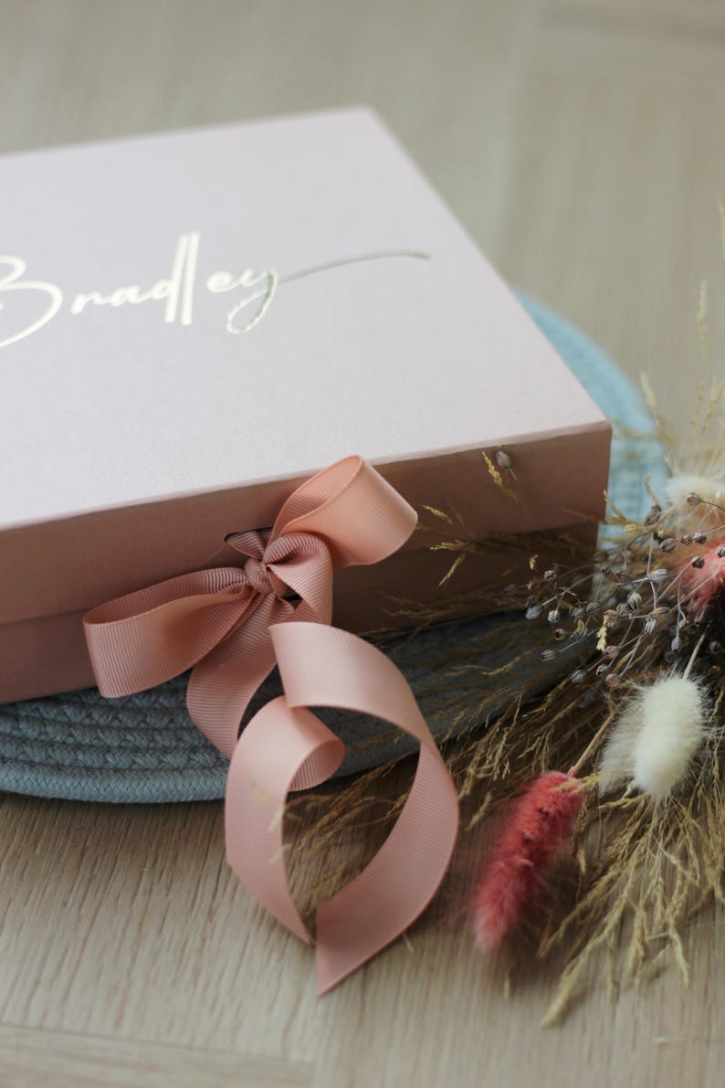 May include: A pink gift box with a gold inscription that reads "Bradley" and a blush pink satin ribbon tied in a bow.