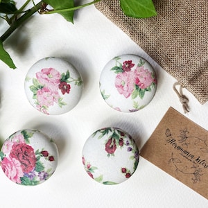 May include: Four white ceramic knobs with a floral pattern featuring pink and red roses, green leaves, and a white background. The knobs are arranged in a square shape on a white surface. The text "Floramana Kitchen" is visible on a brown card.