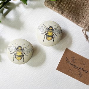 Può includere: Due pomelli rotondi in legno con un design di ape dipinto. Le api sono gialle e nere con ali bianche. I pomelli sono su uno sfondo bianco.